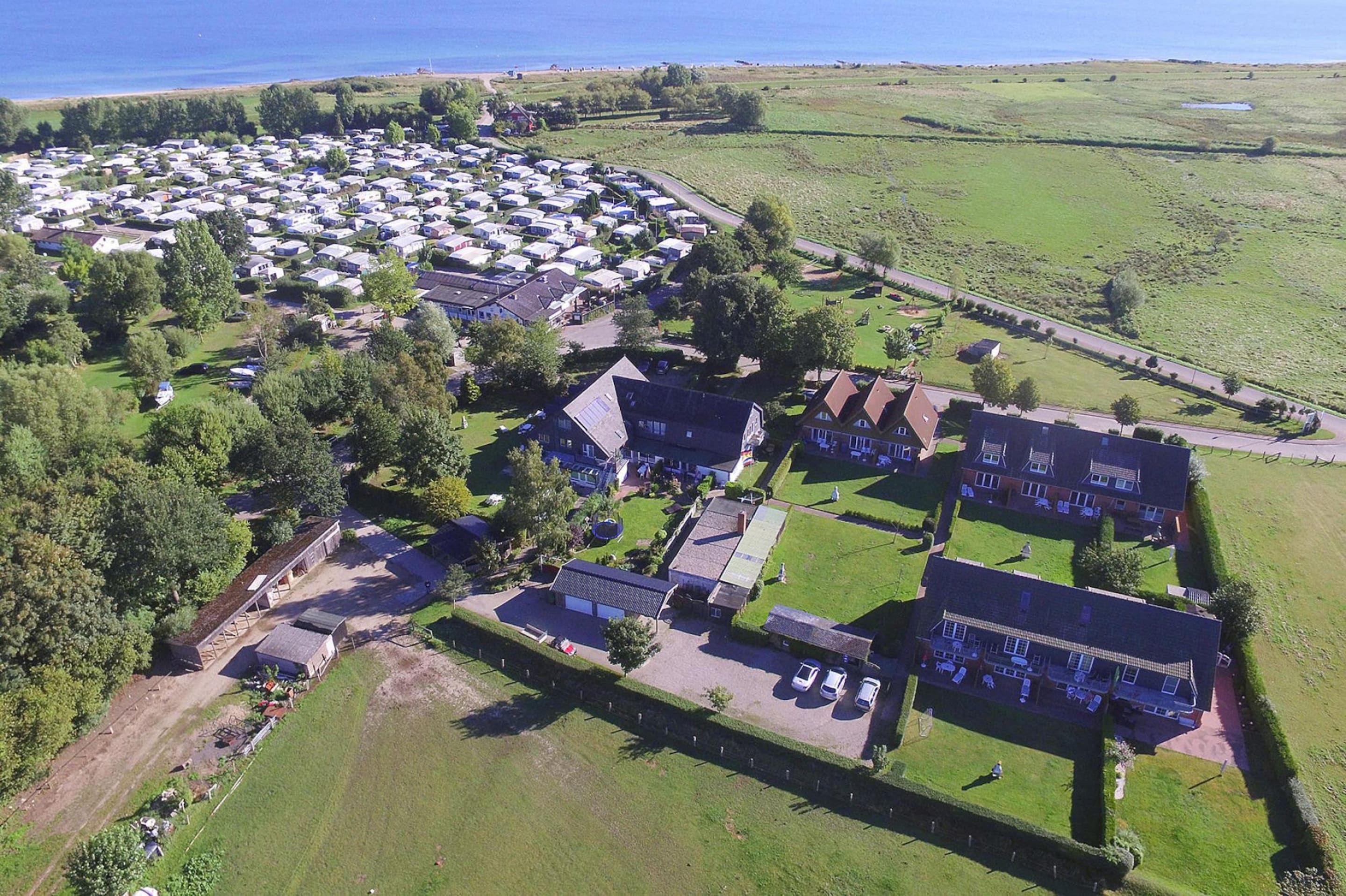 Eine Vogelperspektive auf einen Campingplatz mit mehreren Wohnmobilen und Häusern, umgeben von grünen Feldern und einem Gewässer.