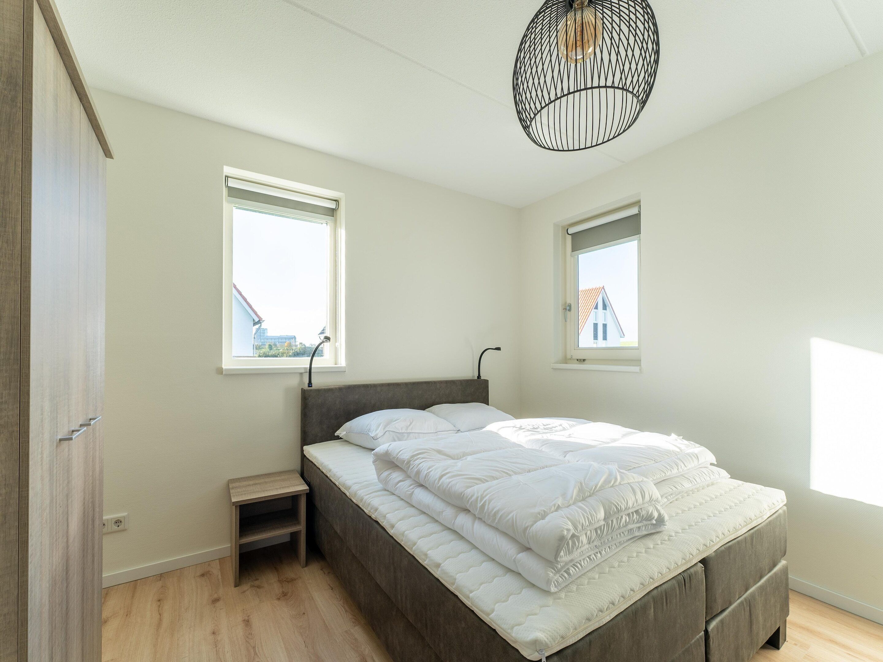 A bedroom with a bed, two windows, a nightstand, and a hanging light fixture.