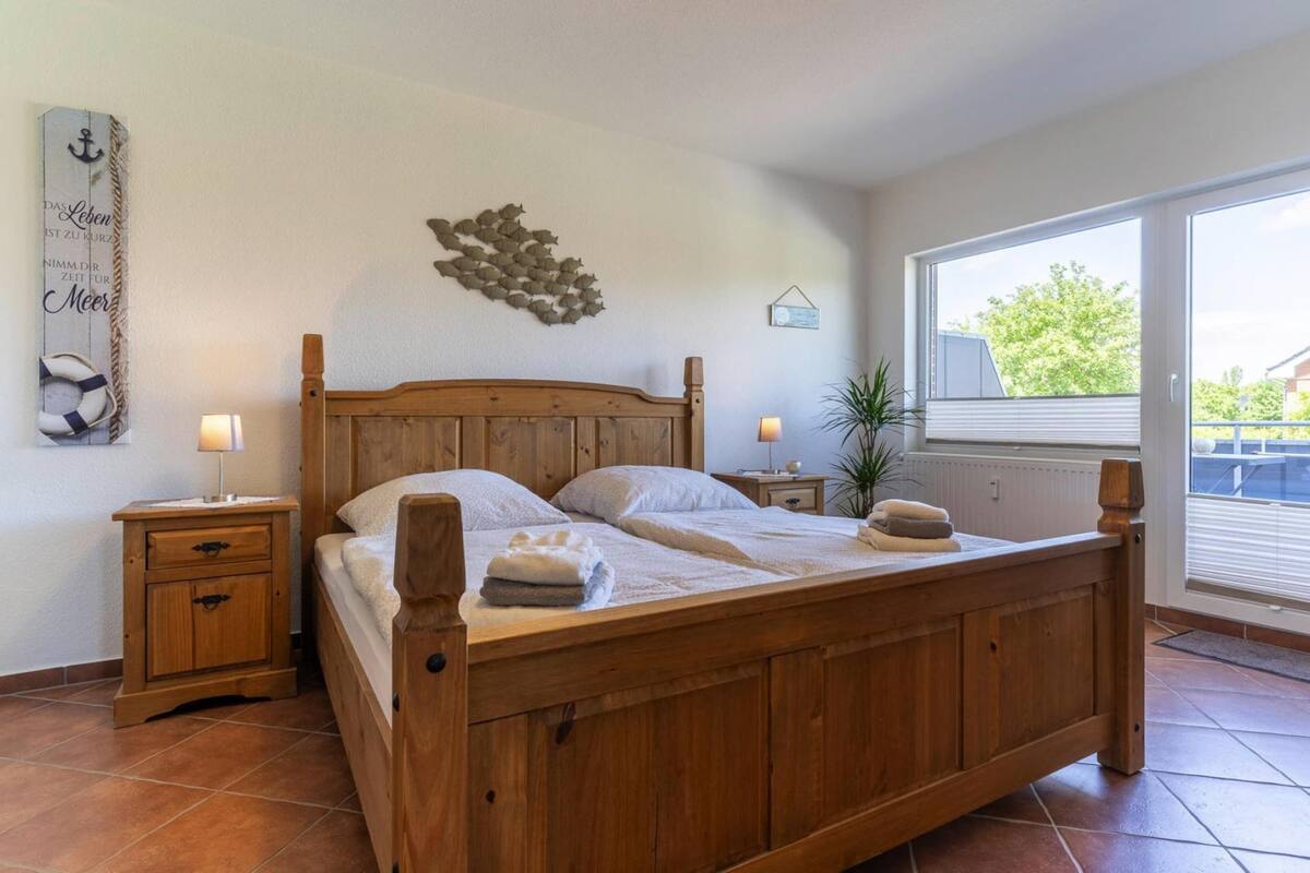 A wooden bed with two pillows, a nightstand with a lamp, a wall decoration, and a window with a view of greenery.