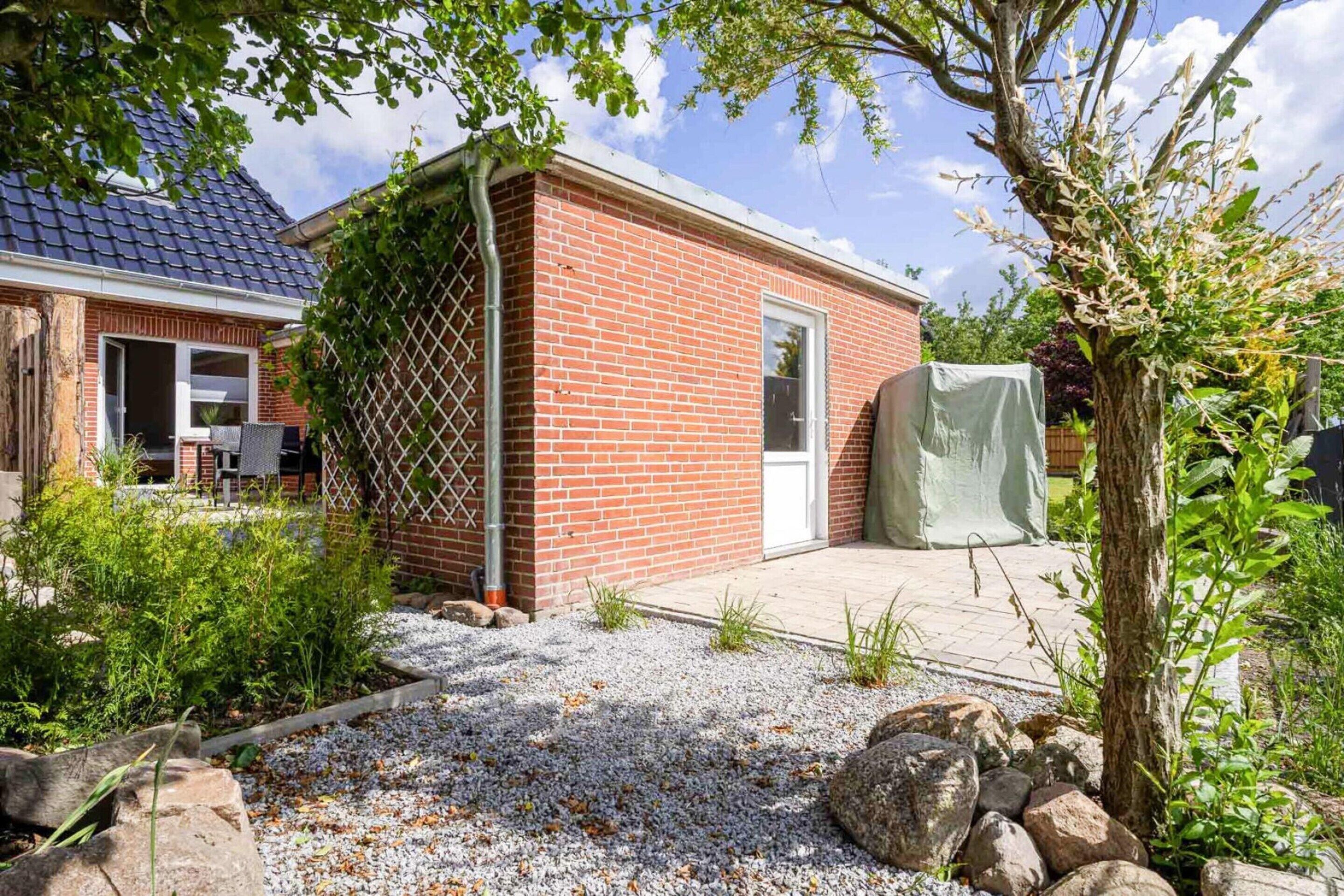 A brick building with a door and window, surrounded by gravel and rocks, with a tree and plants nearby.