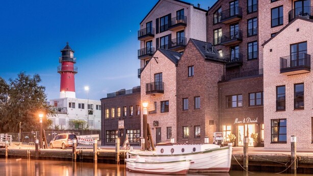 Ett hotell vid vattnet med byggnader i tegel, en båt förtöjd framför och en fyr i bakgrunden.