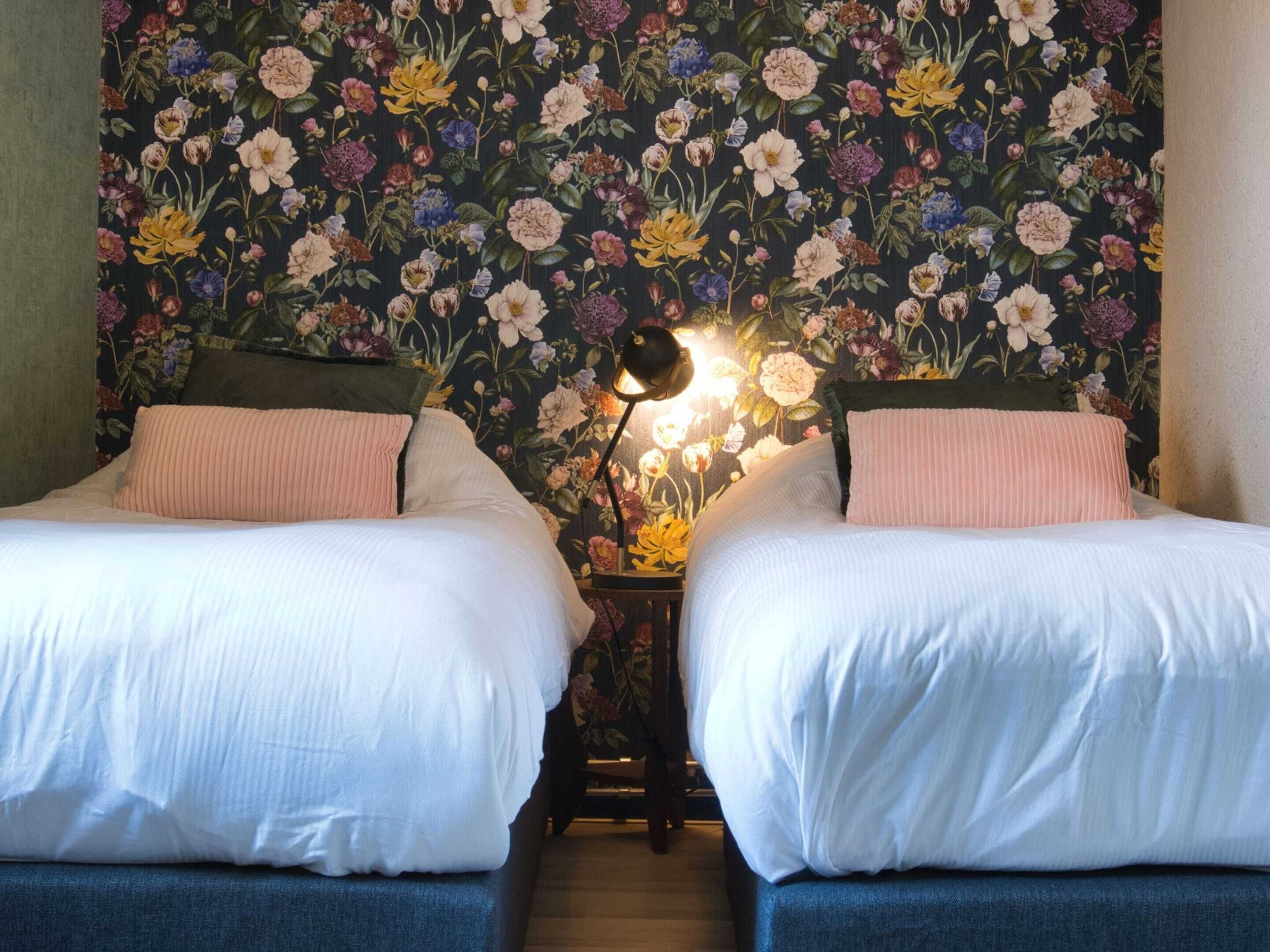 A room with two beds, a dark floral wallpaper, and a nightstand with a lamp.