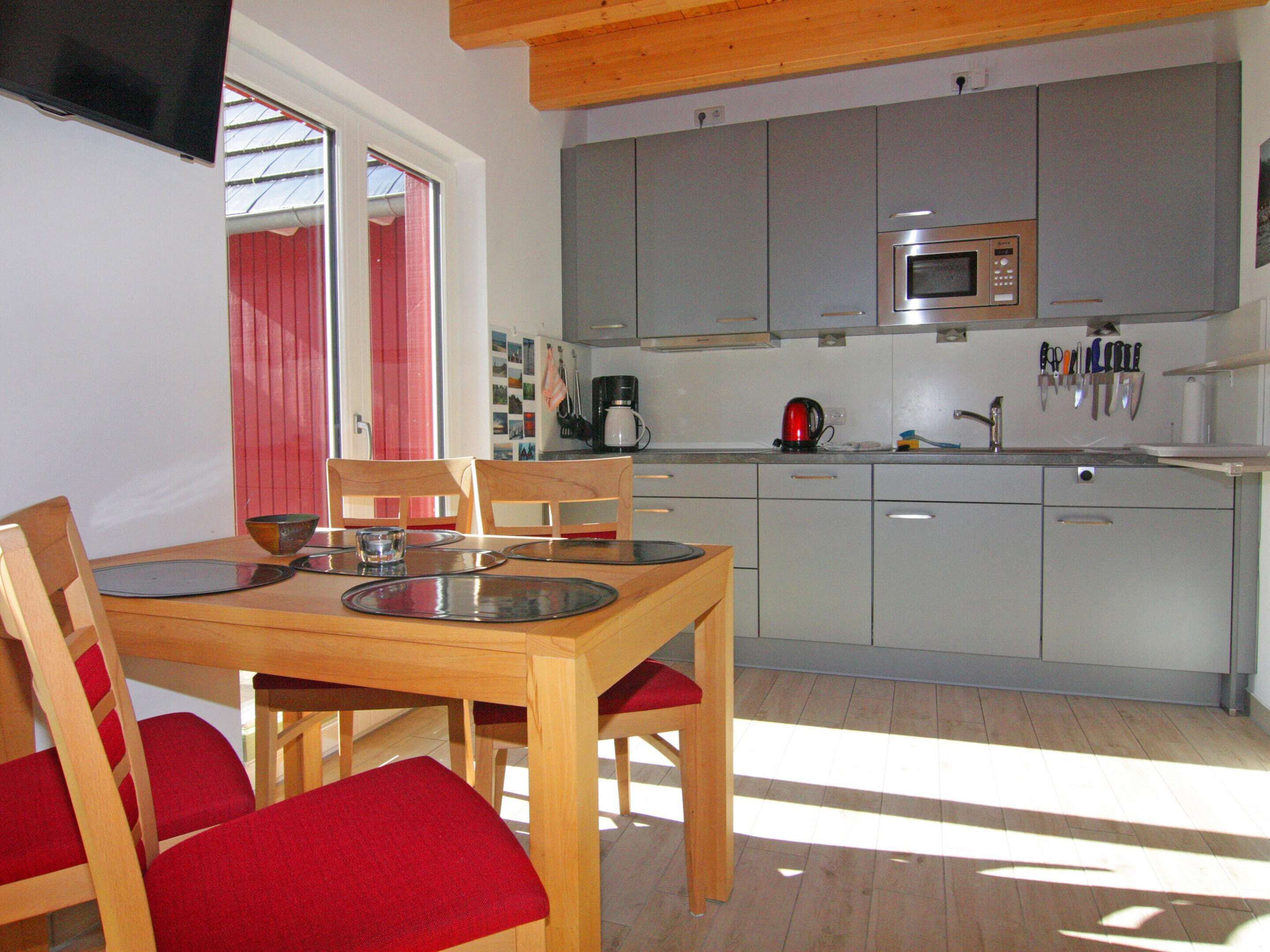 A modern kitchen with grey cabinets, a microwave, and a coffee maker. There is a dining table with chairs and a television mounted on the wall.