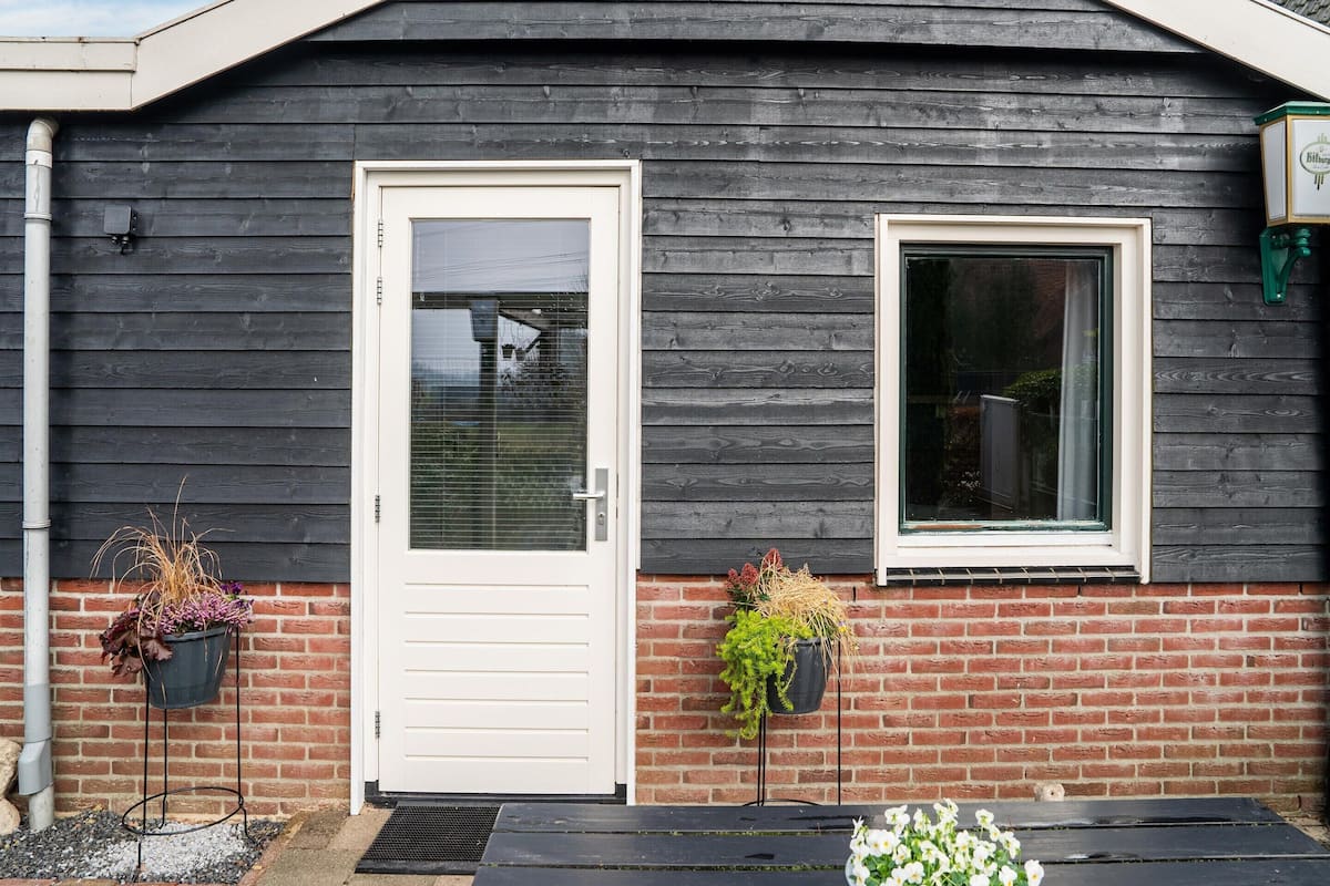 Un bâtiment à l’extérieur en bois foncé, avec une porte blanche et une fenêtre. De chaque côté de la porte, on trouve deux supports en métal noir sur lesquels sont posés des pots de plantes.