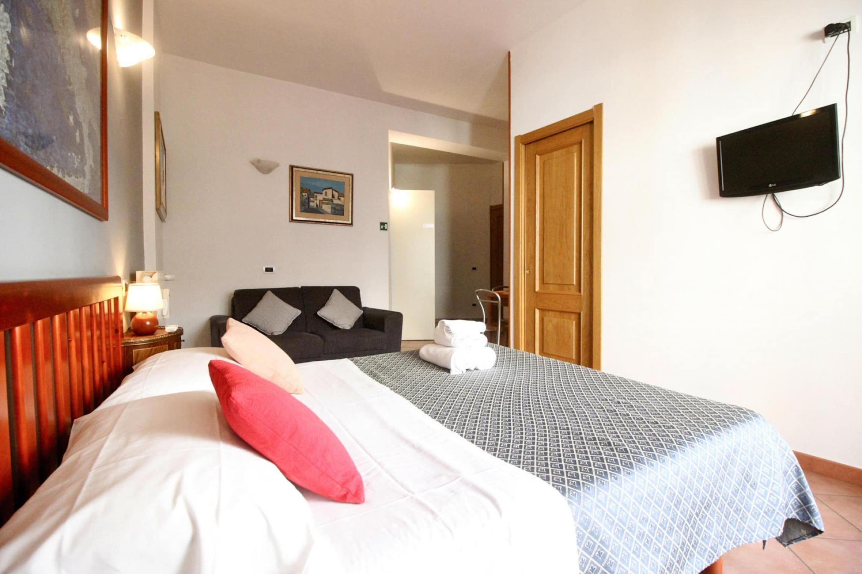 A hotel room with a bed, a nightstand with a lamp, a TV mounted on the wall, and a door leading to another room.