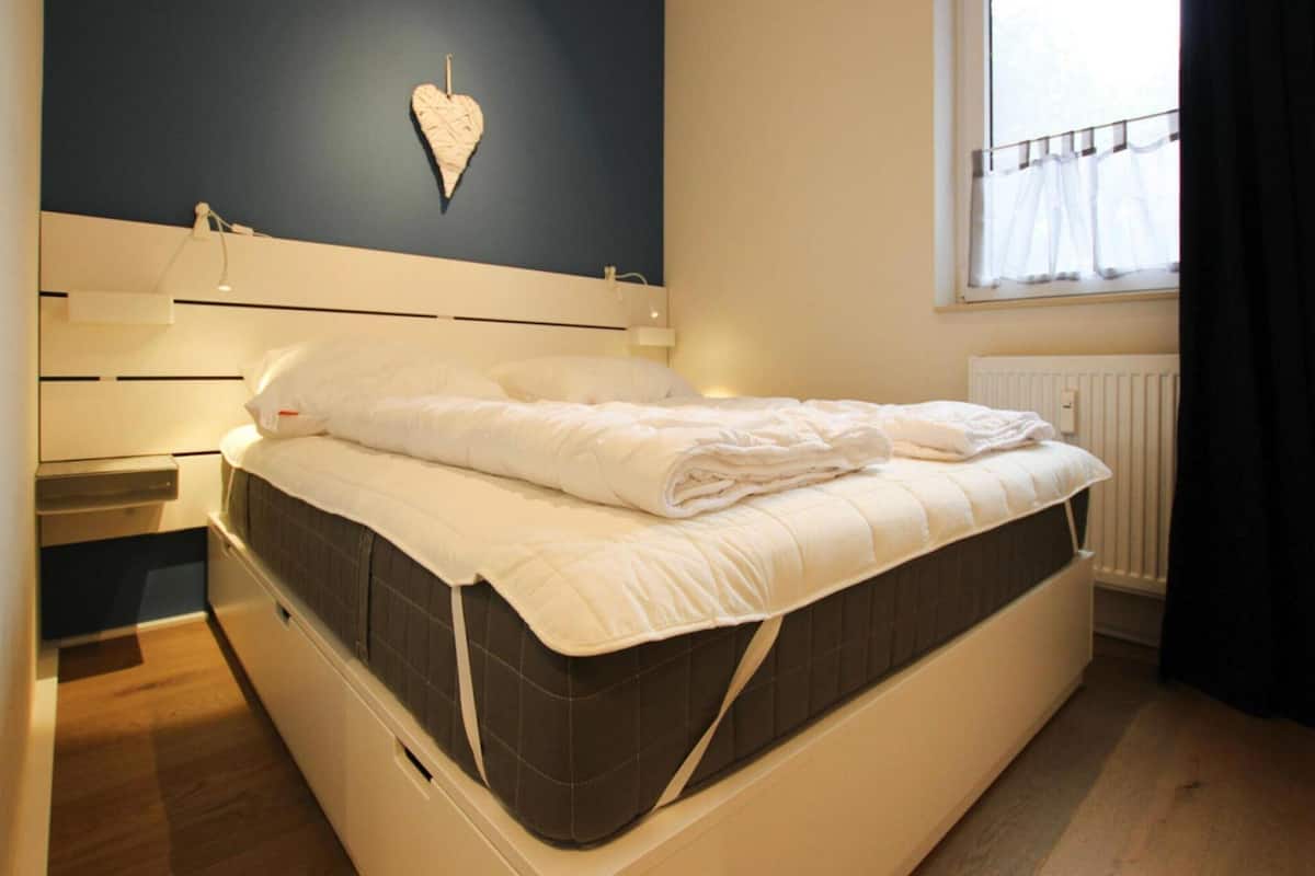 A neatly made bed with a quilted mattress, a bedside table with a lamp, and a heart-shaped decoration on the wall.