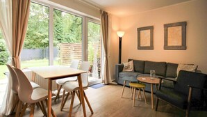 A modern living room with a dining table, chairs, a sofa, and a small coffee table. There are two framed pictures on the wall and a lamp on a side table. - Apartment, St. Peter-ording (St. Peter-Ording)