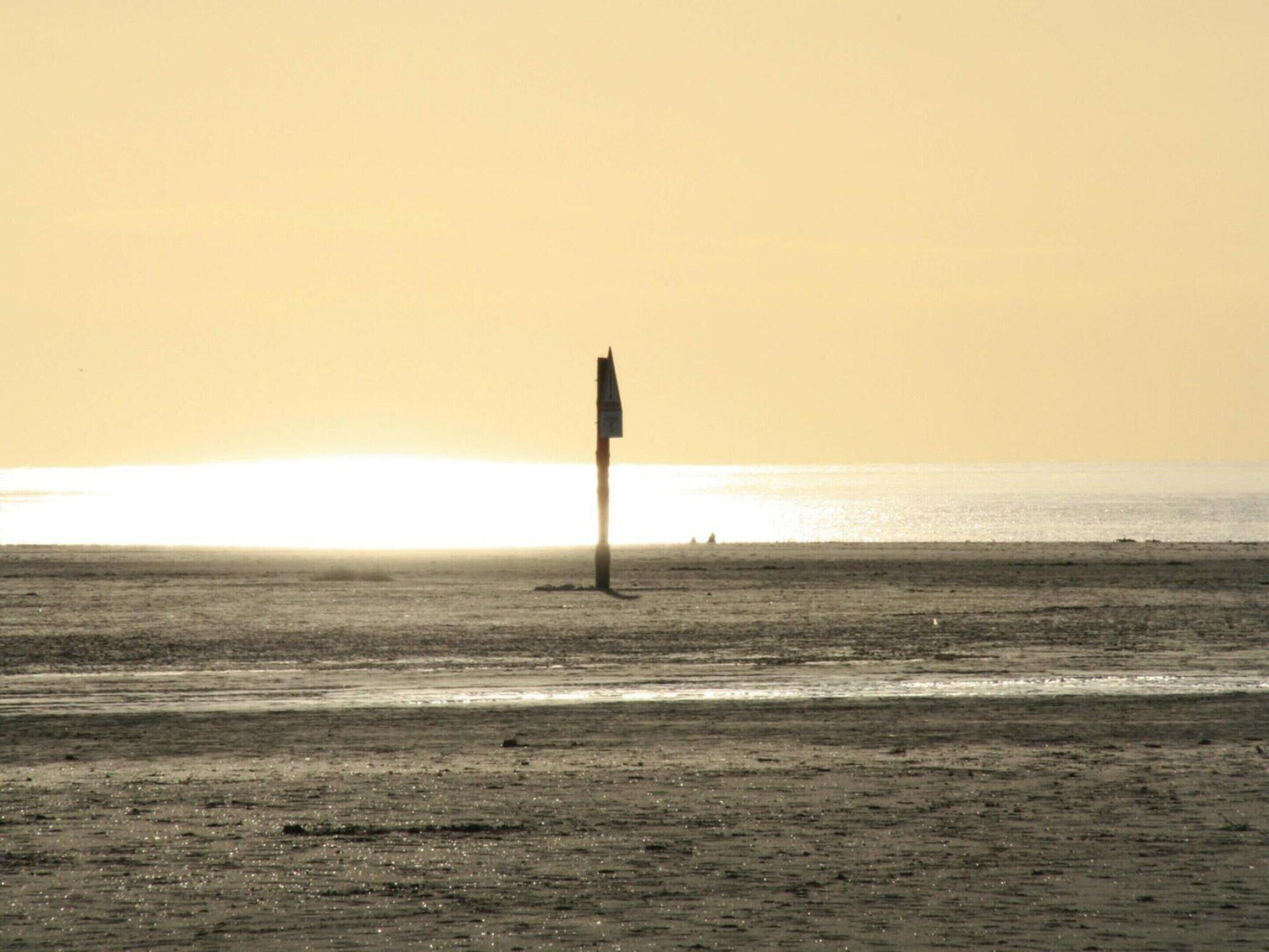 A beach with a solitary flagpole in the distance, a calm sea, and a clear sky.