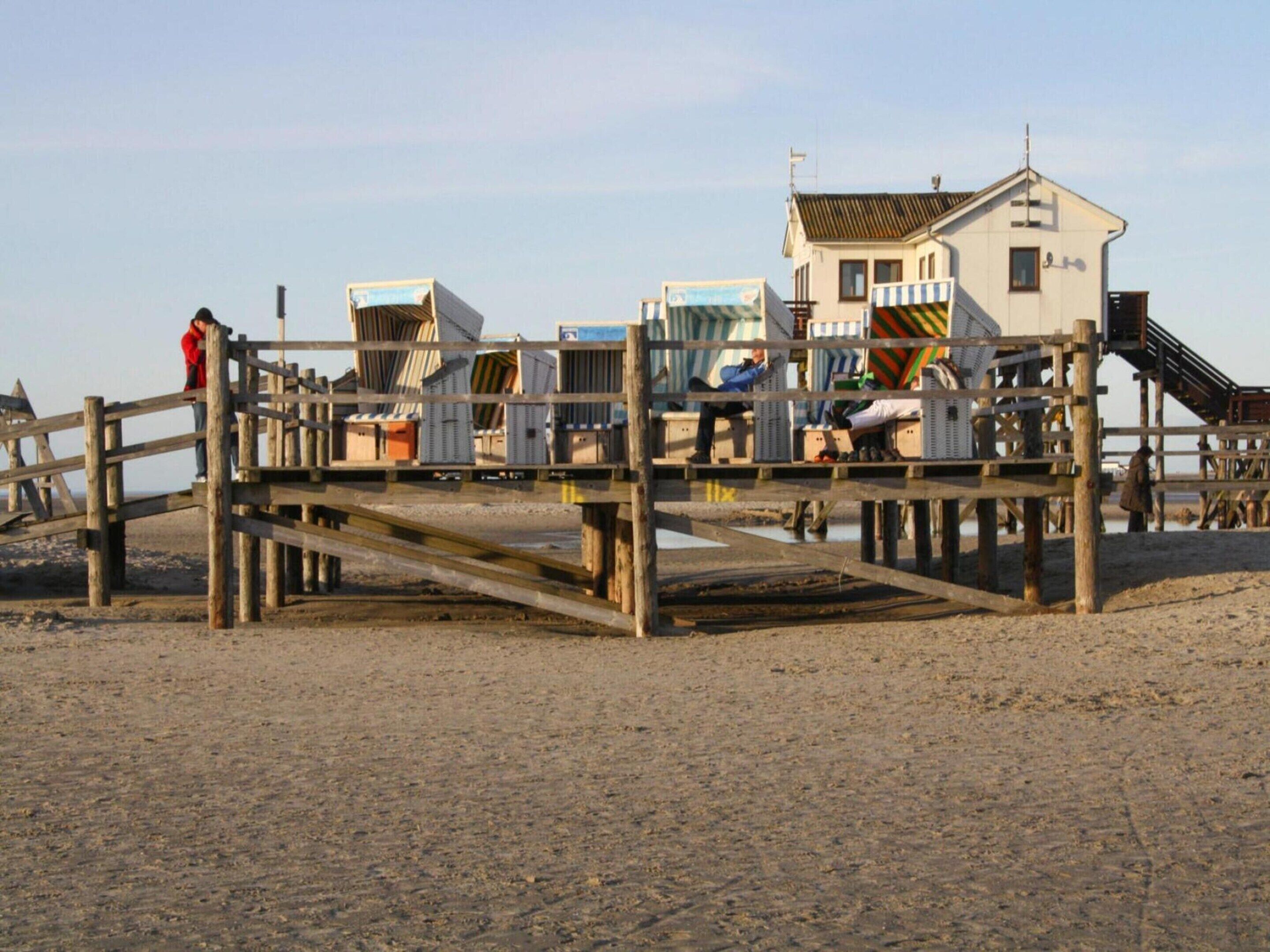 En træbro med en række små bygninger, sandsynligvis en badeanstalt ved stranden, med en trappe, der fører op til en større bygning i baggrunden.