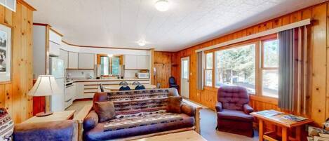 Interior de una cabaña con sofá, sillón, mesa de comedor y zona de cocina.