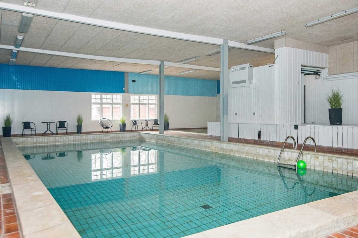 Une piscine intérieure avec des carreaux bleu clair, un bord en briques et un mur blanc avec une section bleue. Des chaises et des tables sont disposées le long du mur, et une plante en pot se trouve à droite.
