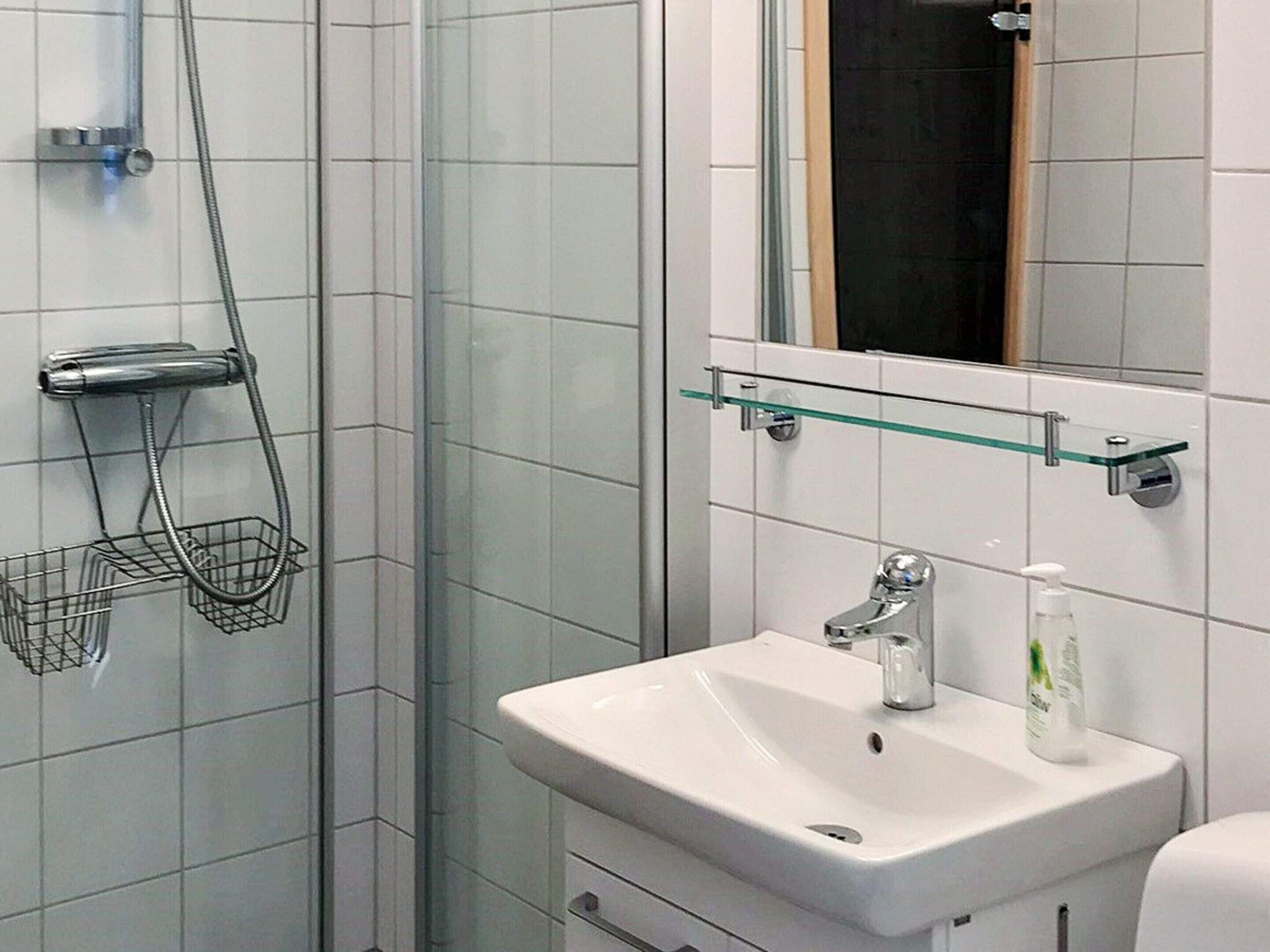 A bathroom with a glass shower enclosure, a white sink with a soap dispenser, and a glass shelf above the sink.