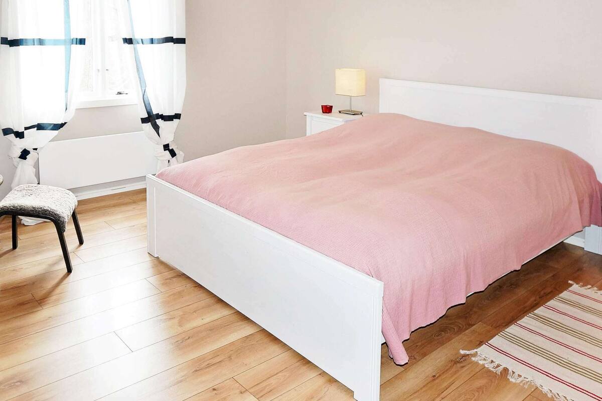 A bedroom with a white bed, a chair, a nightstand with a lamp, and a window with curtains.