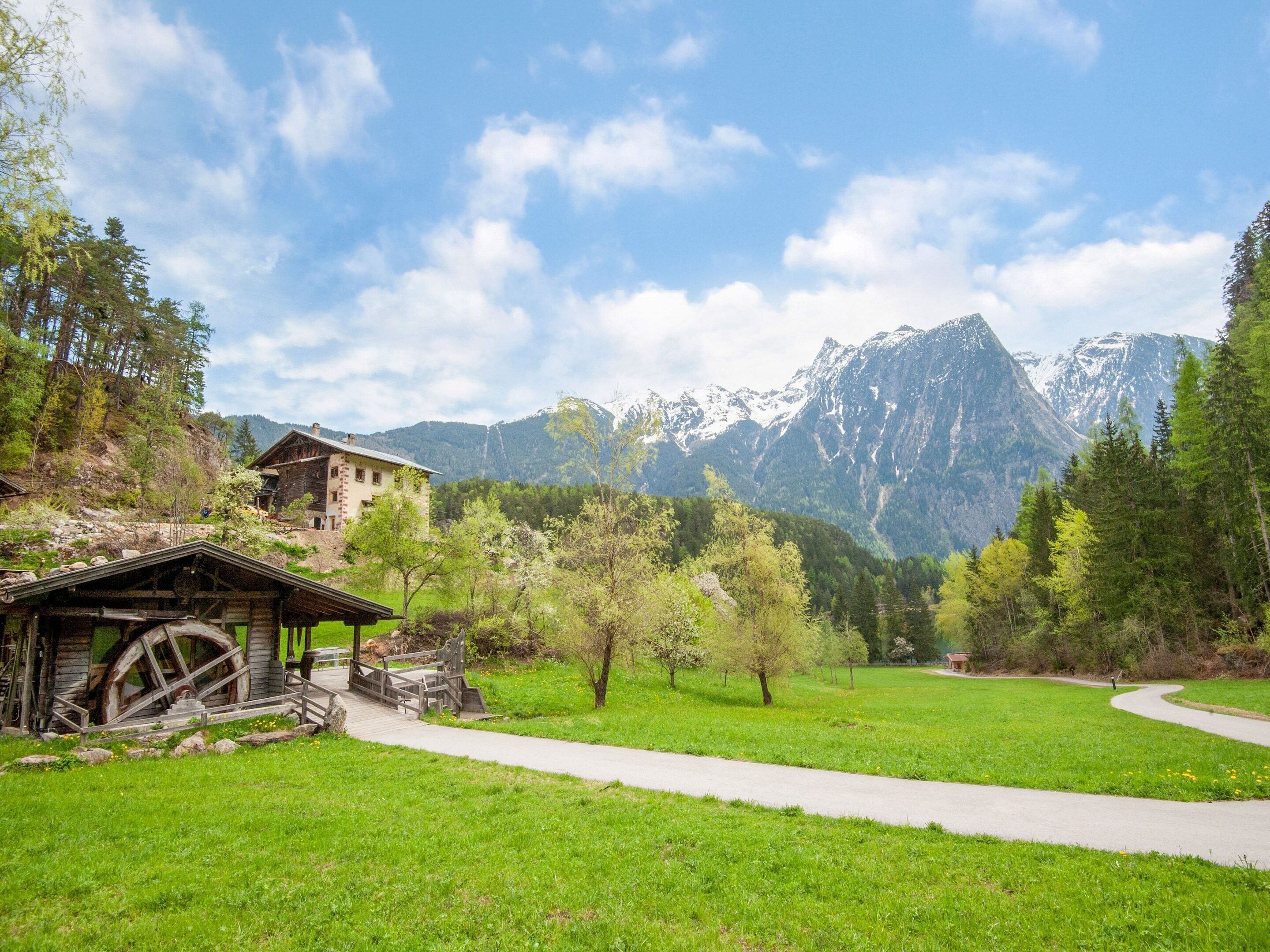 A rustic wooden building with a large water wheel, surrounded by lush green fields and mountains in the background.