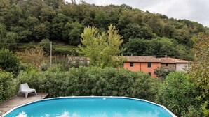A swimming pool with a wooden deck and a white chair, surrounded by lush greenery and a hillside in the background. - Belvilla by OYO Holiday Home in Molina di Quosa (San Giuliano Terme)