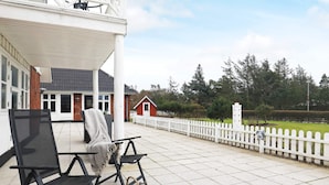 A covered patio area with outdoor seating, a white picket fence, and a view of a red building and trees in the background. - 2 Person Holiday Home in Blavand (Blavand)