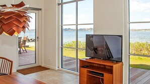 A modern living room with a large TV on a wooden stand, a dining table, and a view of a body of water through large windows. - 5 Person Holiday Home in Farso (Farso)