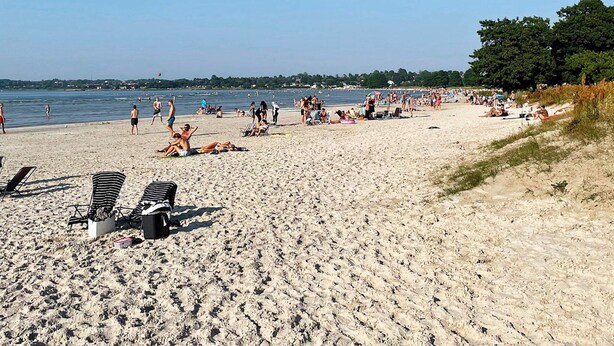 Uma praia arenosa com pessoas tomando sol e nadando, e uma vista clara da água.
