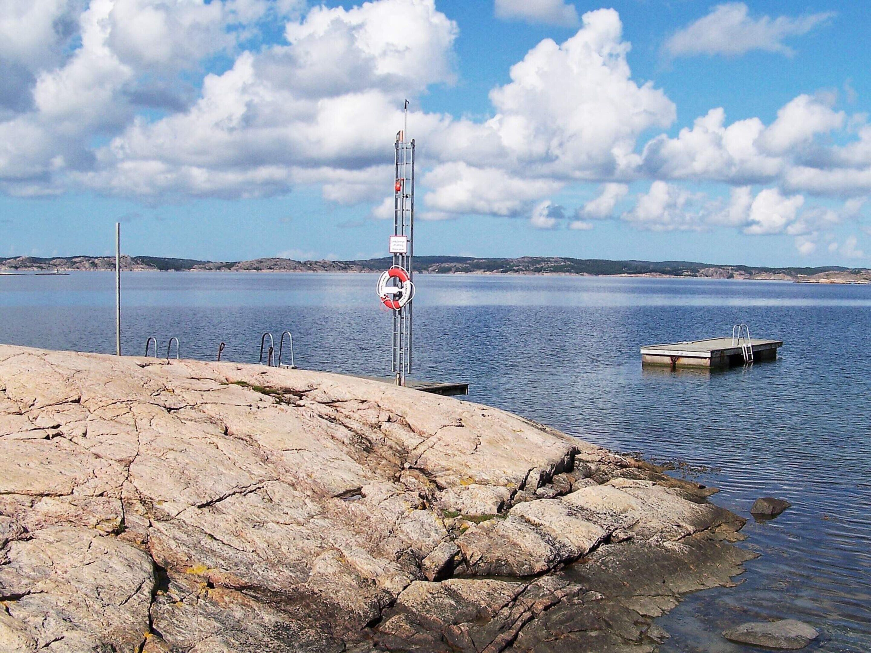 A rocky shoreline with a lifebuoy attached to a pole, a dock, and calm water.