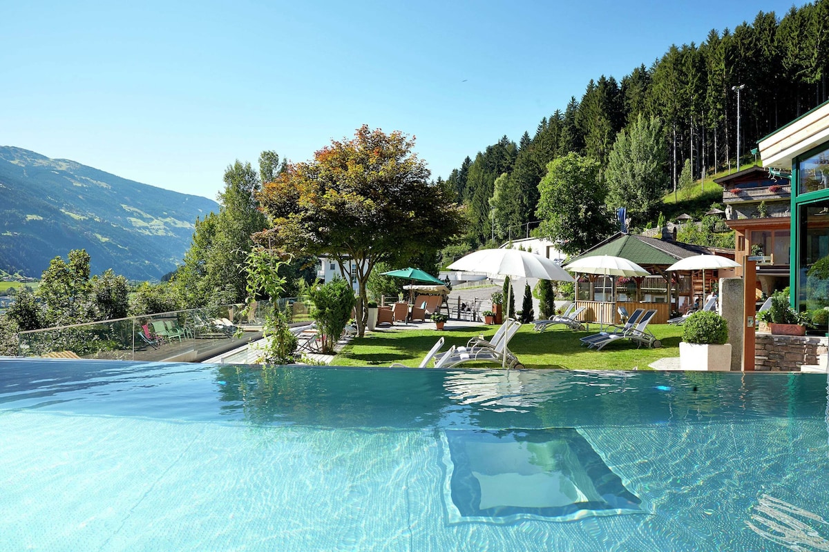 Une piscine aux eaux cristallines, bordée de chaises longues et d’ombrelles, entourée d’un paysage montagneux et d’un bâtiment doté d’un toit végétalisé.