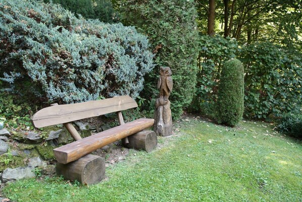 A wooden bench with a backrest and a log seat, positioned on a grassy area with a stone wall and dense shrubbery in the background.