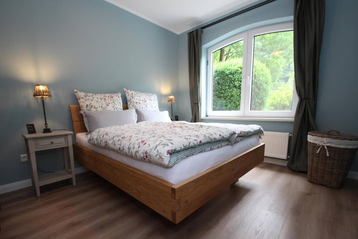 A bedroom with a wooden bed, two bedside tables with lamps, a window with a view of greenery, and a wicker basket.