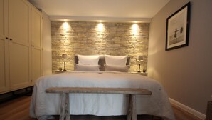 A bedroom with a stone wall, a bed with a wooden bench, and a nightstand with lamps. - Apartment Haus am Berg, Lonau (Herzberg am Harz)