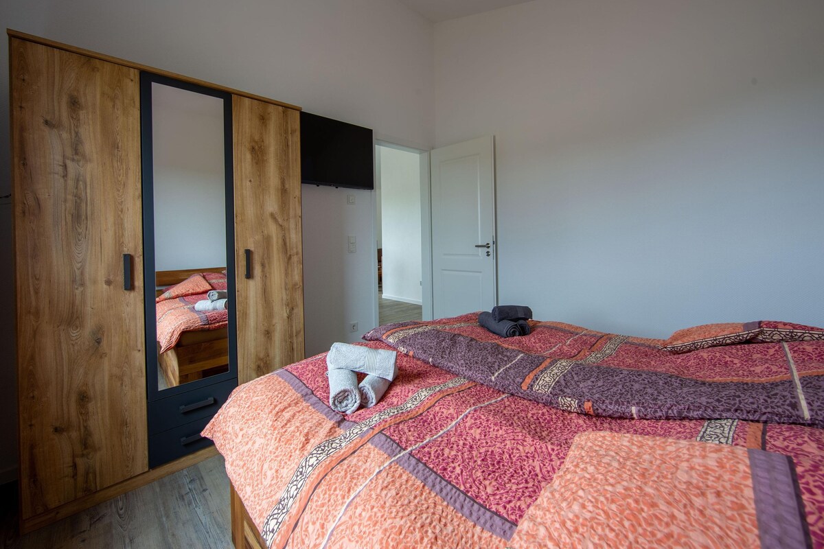 A bedroom with a wooden wardrobe, a bed with patterned bedding, and a mirror reflecting the room.
