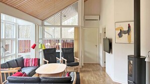 Uma sala de estar moderna com teto de madeira, janelas amplas, um sofá de couro preto, duas poltronas, uma mesa redonda pequena, uma lareira e uma TV fixada na parede.