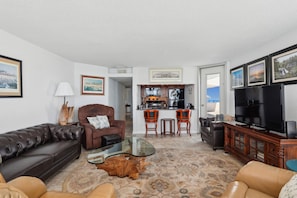 A living room with a brown leather sofa, a glass coffee table, a brown armchair, a TV, and a kitchen area in the background.