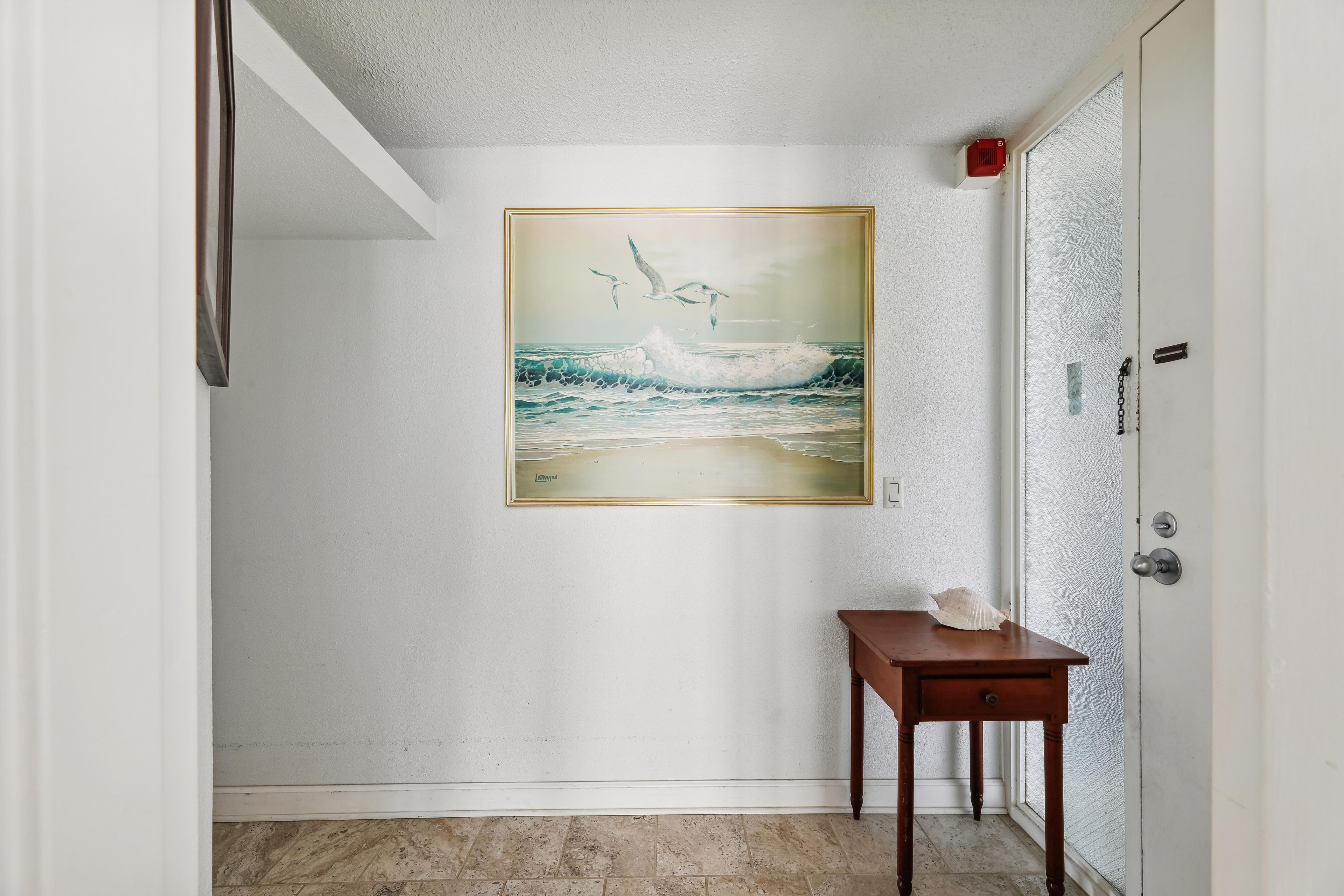 A hallway with a painting of seagulls and waves on the wall, a small wooden table, and a door with a security device.