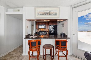A compact kitchen with a bar area, two stools, and a view of the beach through the door.