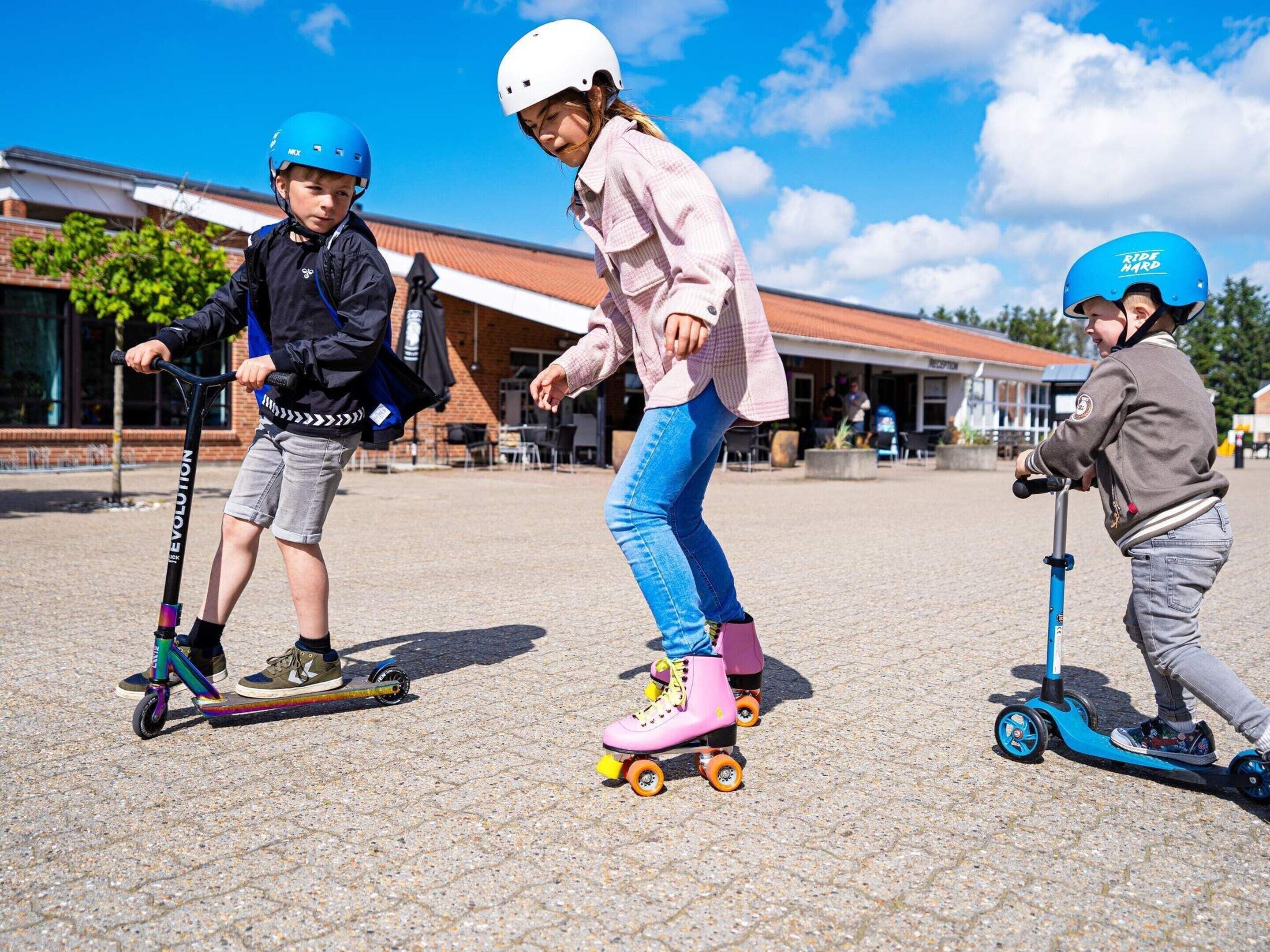 Three children riding scooters in an outdoor area with a building in the background.