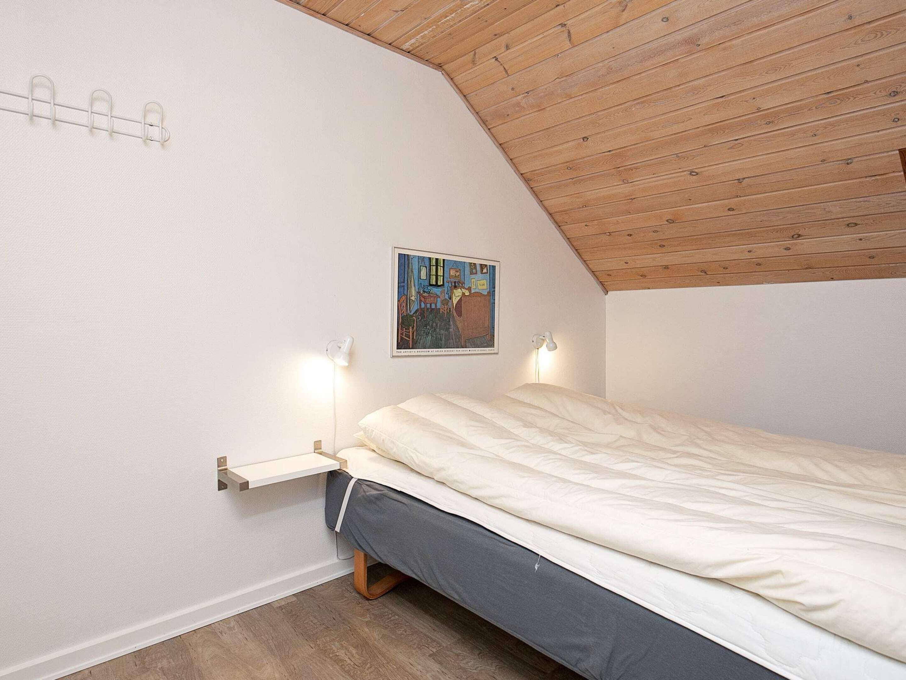 Ein Schlafzimmer mit schrägem Dach, einem Bett mit grauer Matratze und weißer Bettwäsche, einem kleinen Nachttisch mit Lampe und einem gerahmten Bild an der Wand.