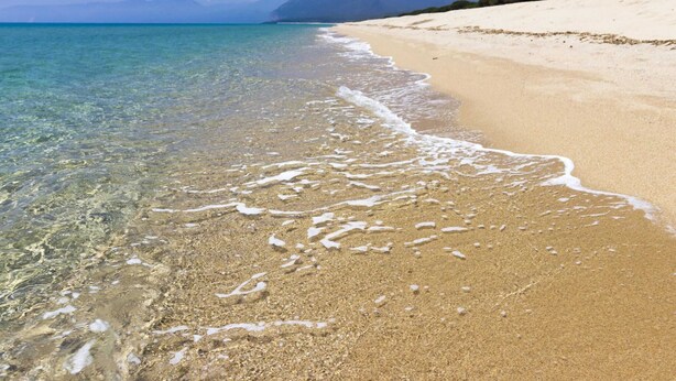 Een strand met helder ondiep water, een zandstrand en een berg op de achtergrond.