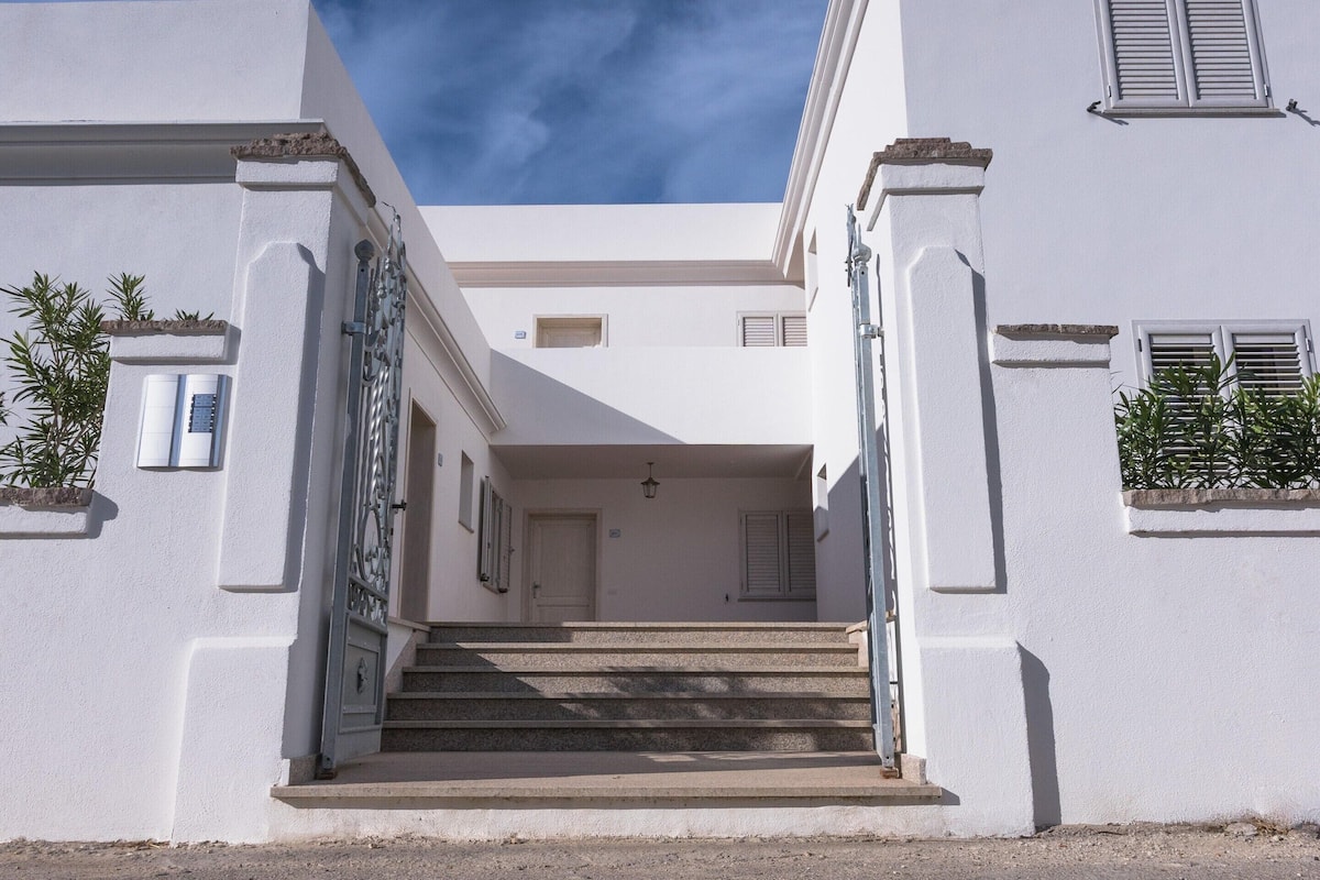 A white building with a central staircase leading to a door, flanked by windows and plants.