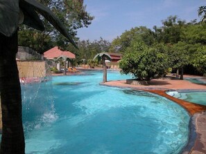 A swimming pool with a curved design, surrounded by lounge chairs and shaded areas. - Aayush Resort Karjat (Panvel)