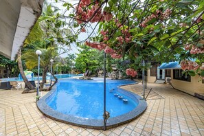 A swimming pool with a tiled deck, surrounded by lush greenery and a stone wall. - Aayush Resort Karjat (Panvel)