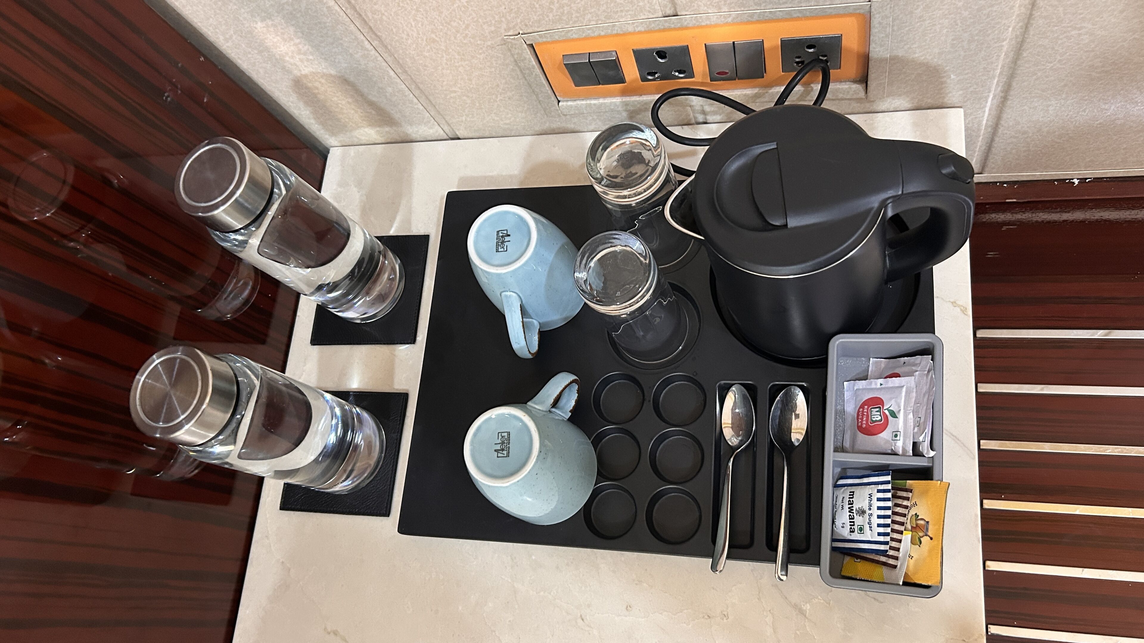 A breakfast setup with a kettle, cups, a teapot, a coffee pot, a muffin tray, a spoon, a knife, and a packet of sugar.
