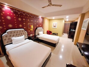 A hotel room with two beds, a ceiling fan, a small table, a red chair, and a TV. - Aayush Resort Karjat (Panvel)