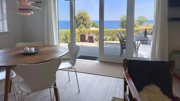 A dining area with a table and chairs, a view of the sea through large glass doors, and a ceiling light fixture. - 4 Person Holiday Home in Gudhjem (Gudhjem)