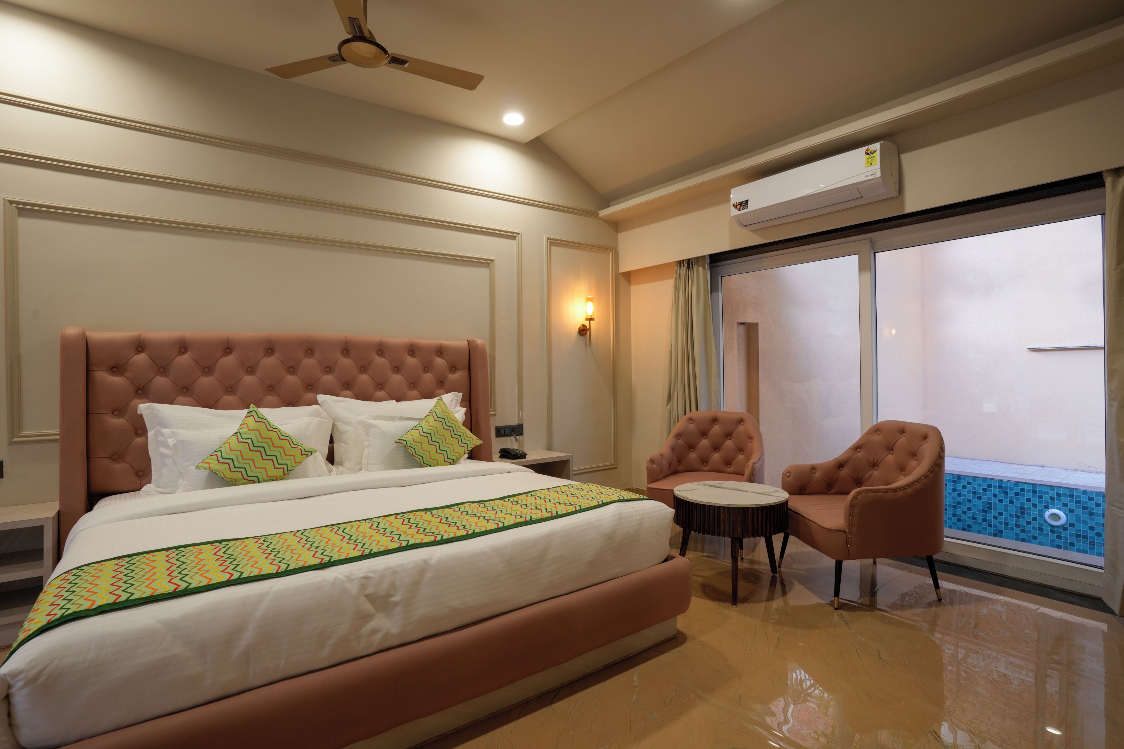 A hotel room with a large bed, a ceiling fan, an air conditioning unit, a small table with two chairs, and a view of a swimming pool through a sliding door.