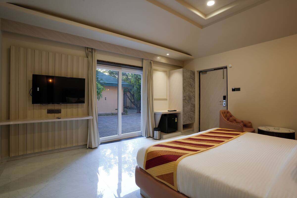A hotel room with a large bed, a flat-screen TV mounted on the wall, a desk with a chair, and a sliding glass door leading to an outdoor area.