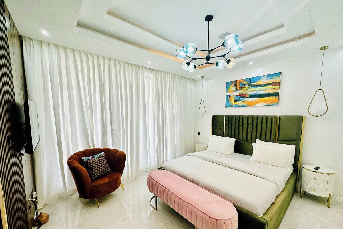 A modern bedroom with a large bed, a bench, a chair, a bedside table, a painting, and a ceiling light fixture.