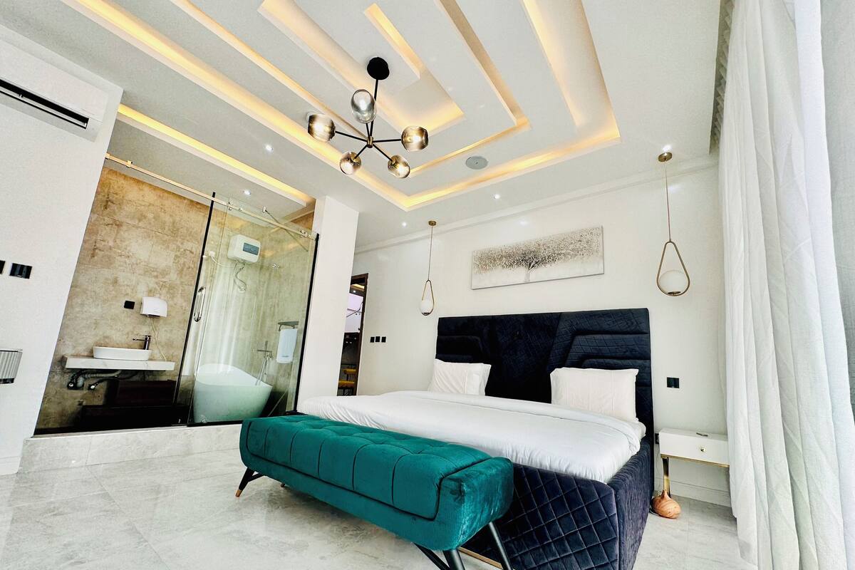 A modern bedroom with a large bed, a teal bench, a dark headboard, and a ceiling with recessed lighting.