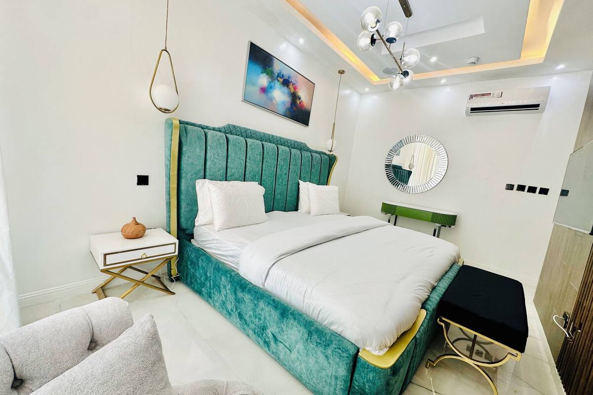 A modern bedroom with a teal upholstered headboard, a white bedspread, and a bedside table with a decorative vase.