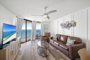 A spacious living room with a large flat-screen TV, a brown leather sofa, a glass coffee table, and a ceiling fan.