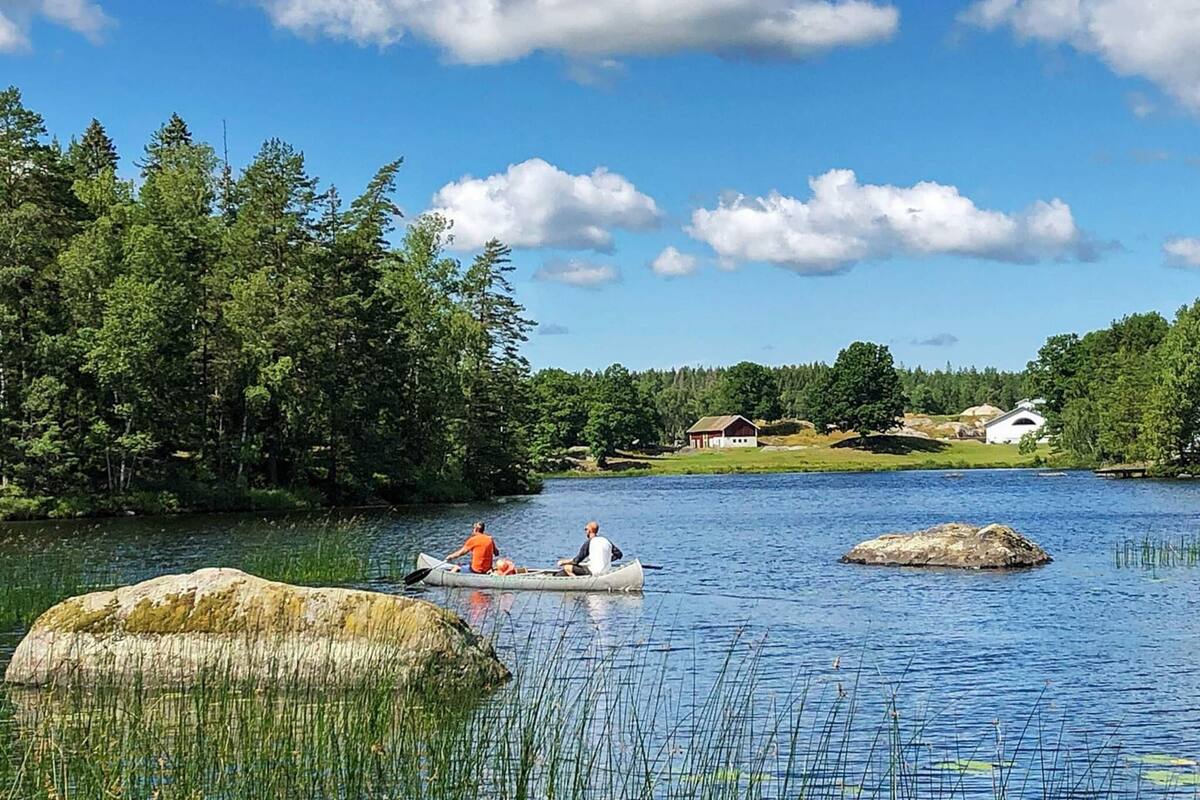 A calm lake with a canoe and two people paddling, surrounded by dense forest and a few buildings in the background.