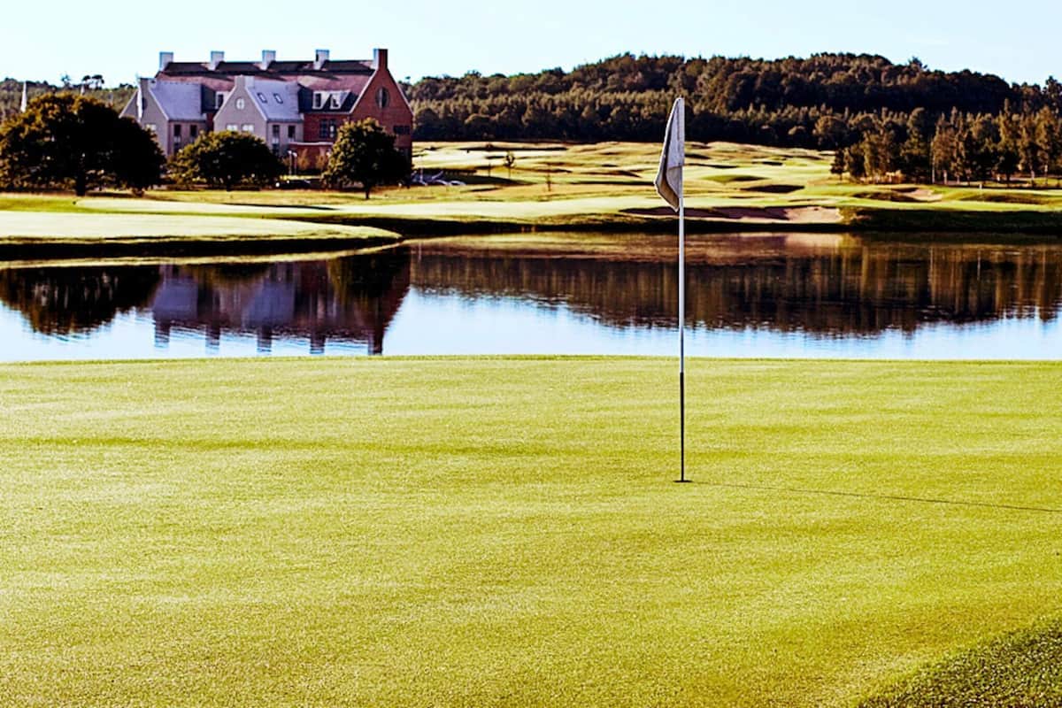 A golf course with a flagstick in the foreground, a pond reflecting the surroundings, and a large house in the background.