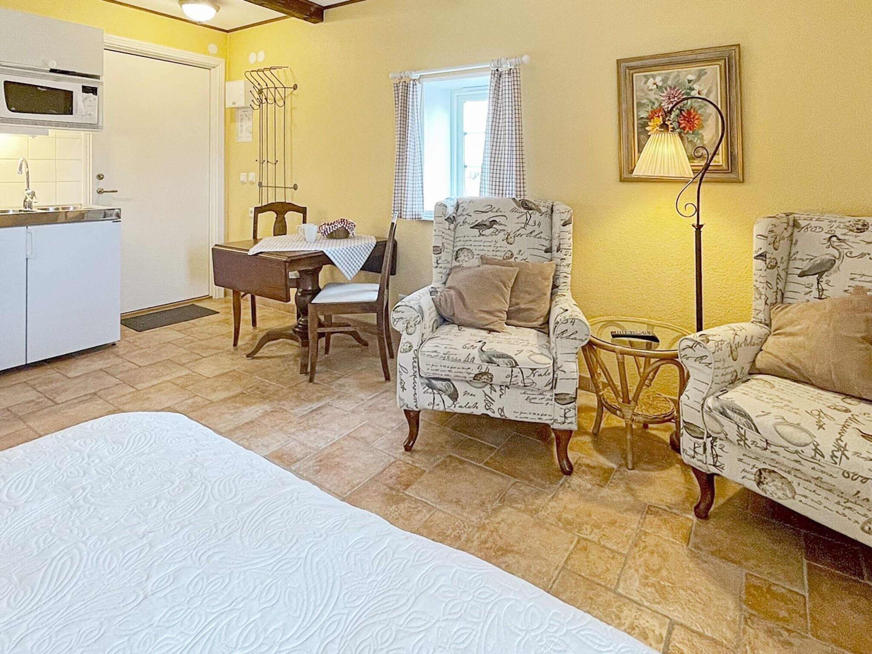 A cozy room with a bed, two armchairs, a dining table with chairs, a kitchen area, and a window with curtains.